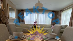 A dining area with a wooden table set for four, surrounded by grey upholstered chairs. A decorative centerpiece with purple flowers and a yellow starburst pattern is placed in the center. The room features a ceiling light fixture and a window with curtains.