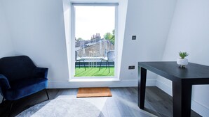 Ein Zimmer mit blauem Sessel, kleinem Tisch mit Pflanze, Fenster mit Blick auf eine Stadtlandschaft und Fußmatte auf dem Boden.