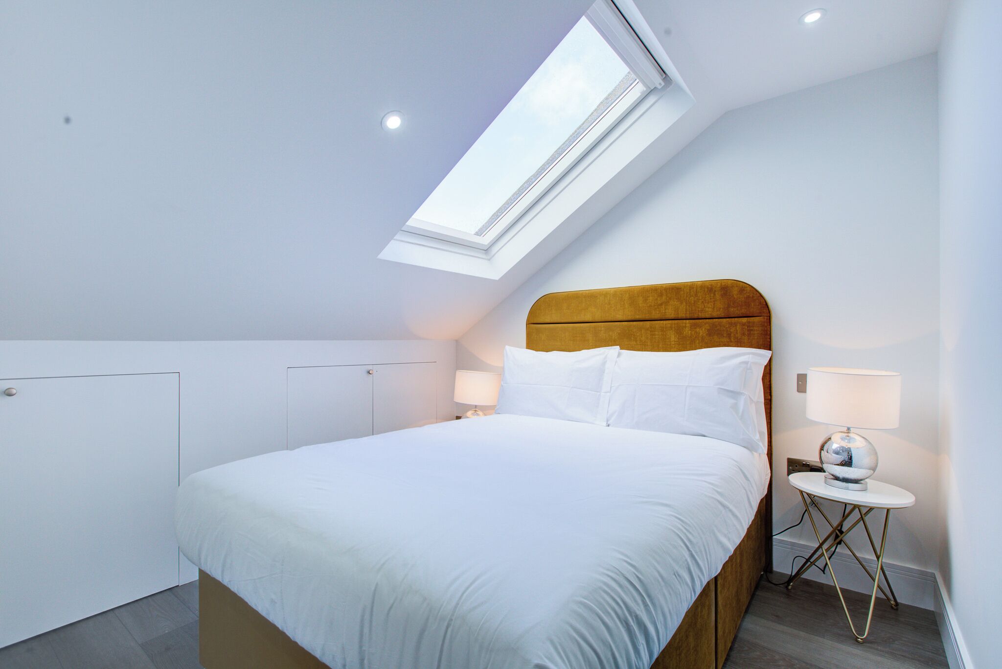 A bedroom with a skylight, a bed with white bedding, a wooden headboard, and a bedside table with a lamp.