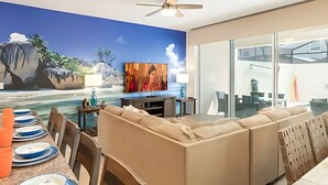 Ett modernt vardagsrum med en stor väggmålning som föreställer en strandscen, en takfläkt, en soffa, en tv och en matplats med tallrikar och glas.