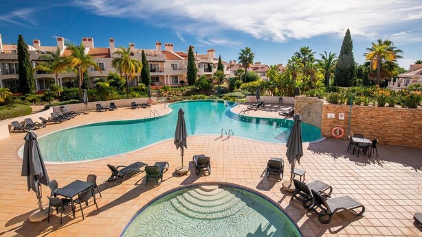 Ein Swimmingpool mit Liegestühlen und Sonnenschirmen, umgeben von Palmen und Gebäuden.