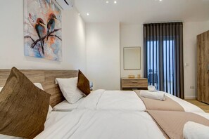 Une chambre d’hôtel moderne avec un grand lit, une tête de lit en bois, un tableau représentant des oiseaux, une table de chevet, un miroir et une vue sur l’extérieur par une fenêtre.