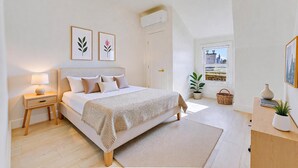 A bedroom with a bed, bedside table, lamp, framed pictures, a window with a city view, and a potted plant. - Spacious 2BR Apartment, 10 min to Time Square! (Weehawken Township)