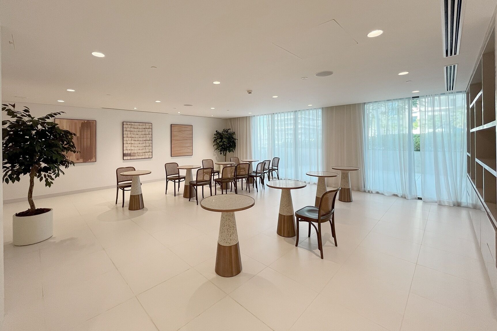 A spacious, well-lit room with multiple round tables and chairs, large windows with sheer curtains, and a potted plant.