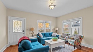 Un salon avec un canapé bleu, une table basse blanche et des fauteuils à motifs. Il y a deux fenêtres avec des cadres blancs et une porte avec un panneau de verre.