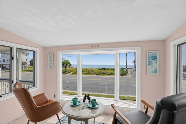Ein Zimmer mit einer großen Schiebeglastür, die einen Blick auf die Straße und das Meer freigibt. Zwei Stühle und ein kleiner Tisch mit Tasse und Untertasse sind ebenfalls vorhanden.