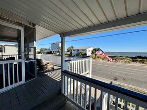 Eine hölzerne Veranda mit Geländer, einem Korbstuhl und Blick auf das Meer und den Strand.