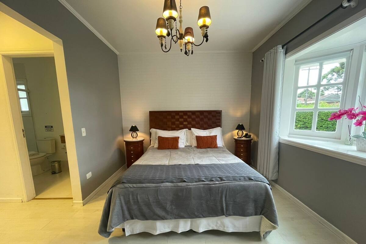 A bedroom with a large bed, two bedside tables, a chandelier, a window with a view, and a door leading to a bathroom.