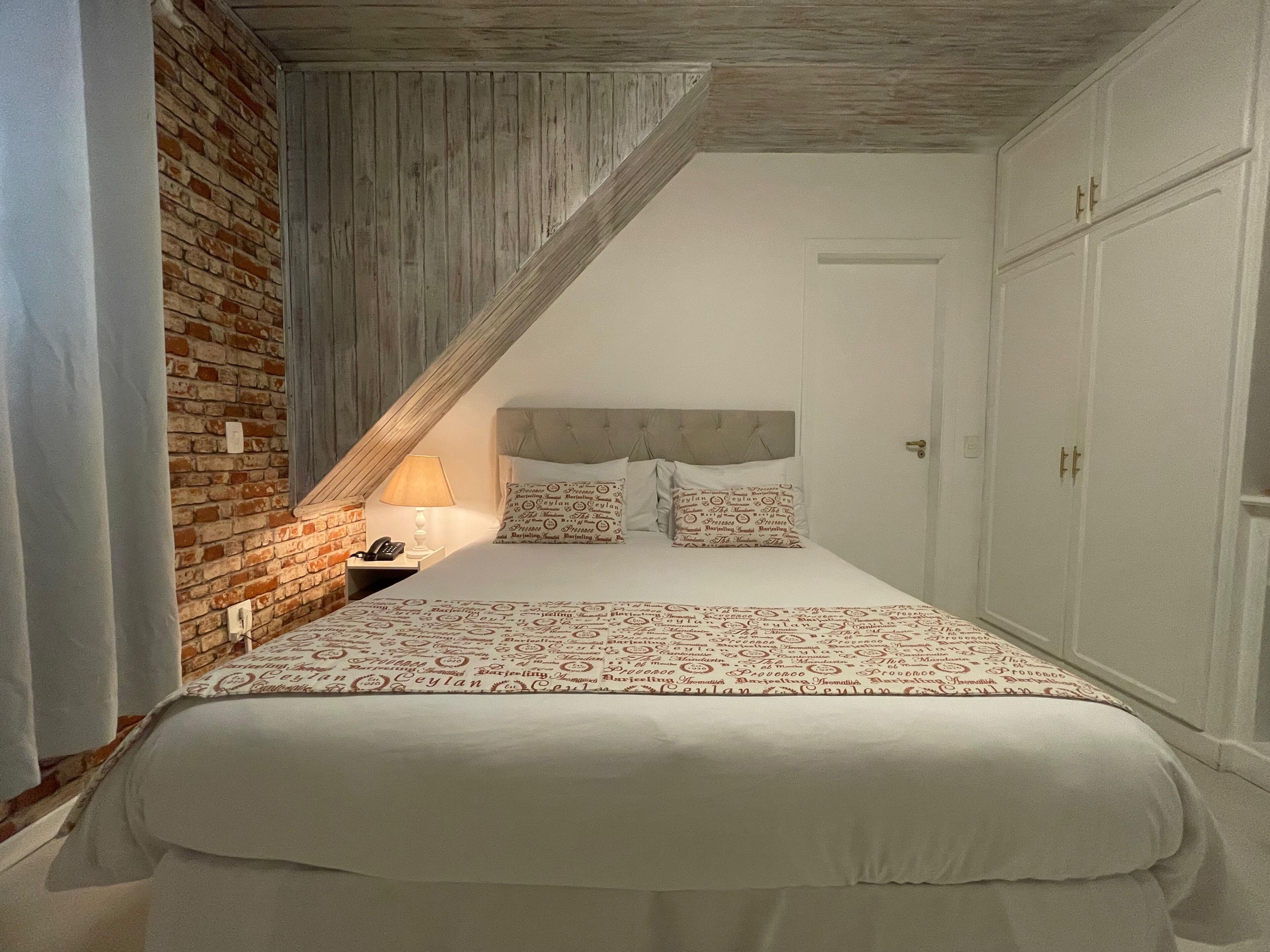 Quarto com teto inclinado, parede de tijolos, cama com roupa de cama estampada, mesa de cabeceira com abajur e guarda-roupa.
