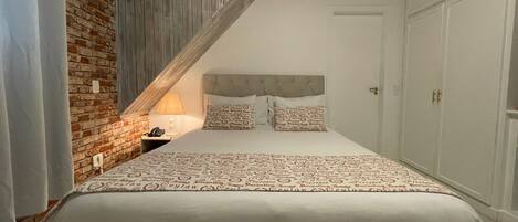 Quarto com teto inclinado, parede de tijolos, cama com roupa de cama estampada, mesa de cabeceira com abajur e guarda-roupa.