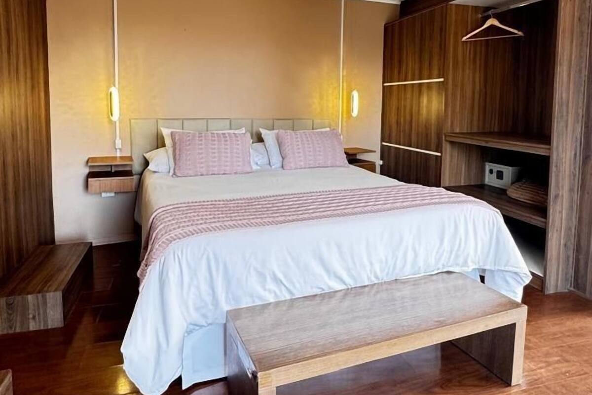 A modern bedroom with a wooden floor, a bed with a white and pink bedspread, a wooden bench, and a wooden wardrobe with a hanging rack.