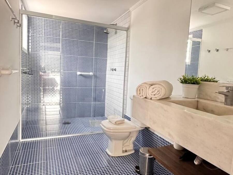 Un baño moderno con ducha revestida en azul, un inodoro blanco y un tocador color beige con espejo.