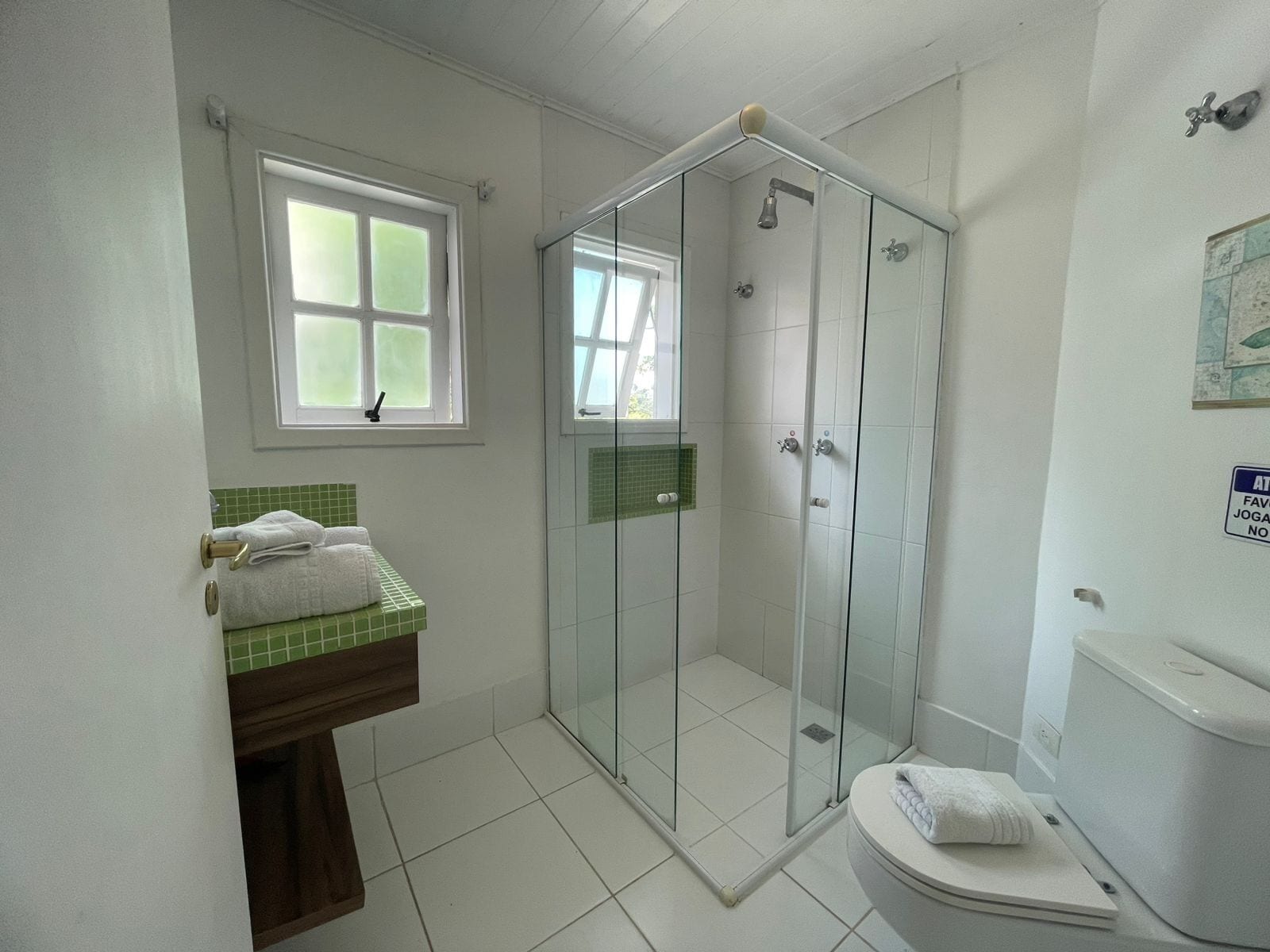 A bathroom with a glass-enclosed shower, a toilet, a sink with a green tiled backsplash, and a window with frosted glass.