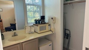 A bathroom with a sink, mirror, and a window with blinds. There is a coffee maker, a hairdryer, and a small refrigerator. - Blue Lagoon Hideaway (Oceano)