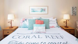 A bed with a coral reef themed bedspread, two bedside lamps, and a framed picture on the wall. - After Dune Delight (Oceano)