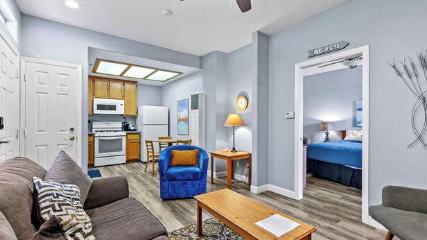 A compact living space with a kitchenette, a sofa, a coffee table, and a bedroom with a bed and a 'BEACH' sign. - The Seaside Sunshine (Oceano)