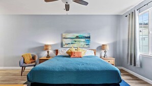 A bedroom with a large bed, two bedside tables with lamps, a chair, and a painting on the wall. - The Seaside Sunshine (Oceano)