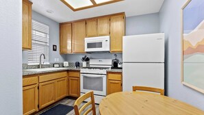 A compact kitchen with wooden cabinets, a microwave, a stove, a refrigerator, and a dining table with chairs. - The Seaside Sunshine (Oceano)
