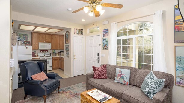 Un salon avec un canapé, un fauteuil et une table basse, une cuisine avec des meubles en bois, un ventilateur de plafond et une grande fenêtre avec des rideaux.