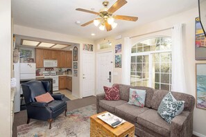 Ein Wohnzimmer mit einer Couch, einem Sessel und einem Couchtisch, sowie eine Küche mit Holzausbau, einem Deckenventilator und einem großen Fenster mit Vorhängen.
