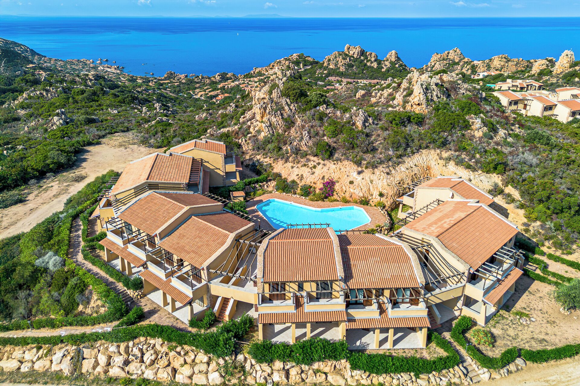 An aerial view of a resort with multiple buildings, a central swimming pool, and a rocky landscape with a view of the sea.