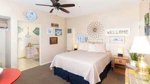 A bedroom with a bed, a ceiling fan, a bedside table with a lamp, a wall clock, and a welcome sign.
