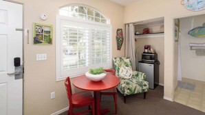A compact room with a dining table, red chairs, a small kitchen area with a microwave, and a window with blinds.