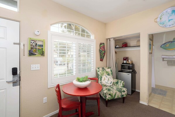 A compact room with a dining table, red chairs, a small kitchen area with a microwave, and a window with blinds. - Waves and Whimsy (Oceano)