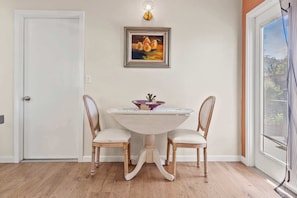 A dining area with a white table and two chairs, a framed painting above the table, and a window with blinds to the right. - The Bay Escape (Baywood-Los Osos)