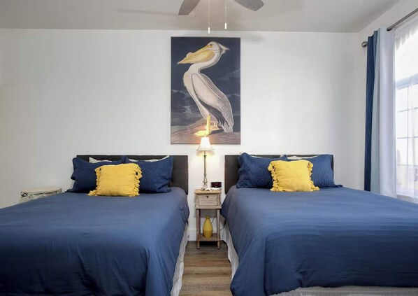 A room with two beds, each with blue bedding and yellow pillows. There is a painting of a pelican on the wall, a bedside table with a lamp, and a window with curtains. - The Seaside Sanctuary (Oceano)