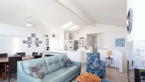 Uma sala moderna com um sofá azul claro, uma poltrona estampada e uma mesa de centro. Há uma área de jantar com mesa e cadeiras de madeira, e uma cozinha ao fundo.