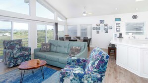 Uma sala de estar ampla, com sofá, poltronas e mesa de centro. Há uma área de jantar com mesa e cadeiras, e uma cozinha com bancada e armários.