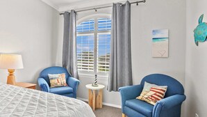 A bedroom with a bed, two blue armchairs, a small round table with a lamp, a window with curtains, and a wall art piece. - Coastal Cutie (Oceano)