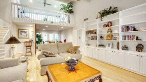 A spacious living room with a high ceiling, a balcony, and a large bookshelf filled with books and decorative items. - Woodlands Escape: 3BR Texas Retreat w/ Forest View (The Woodlands)