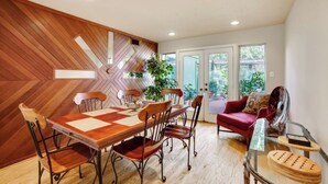 A dining area with a wooden herringbone wall, a large wooden table, and six chairs. There is a glass coffee table with a red cushion and a decorative item on it. - Woodlands Escape: 3BR Texas Retreat w/ Forest View (The Woodlands)