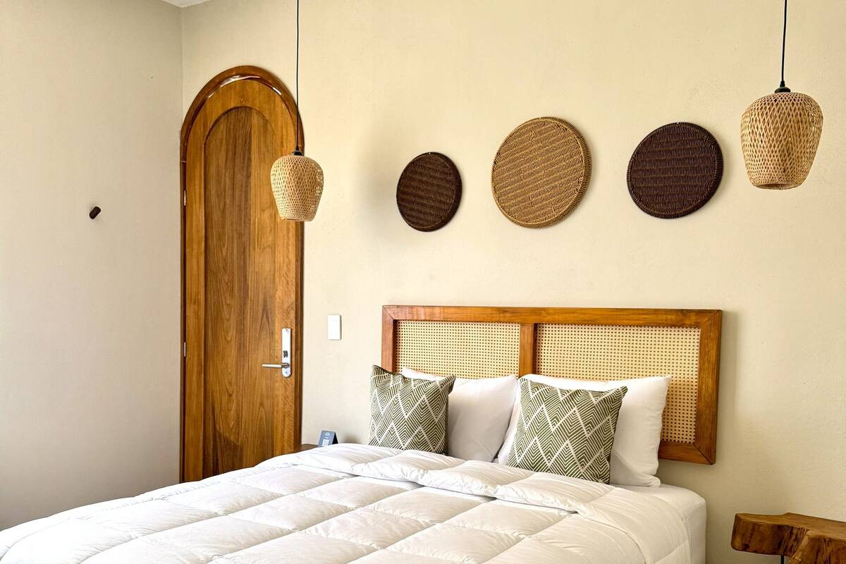 A bedroom with a wooden bed frame, a white quilted bedspread, and a headboard with woven panels. There are four woven wall hangings above the bed and a wooden door to the left.