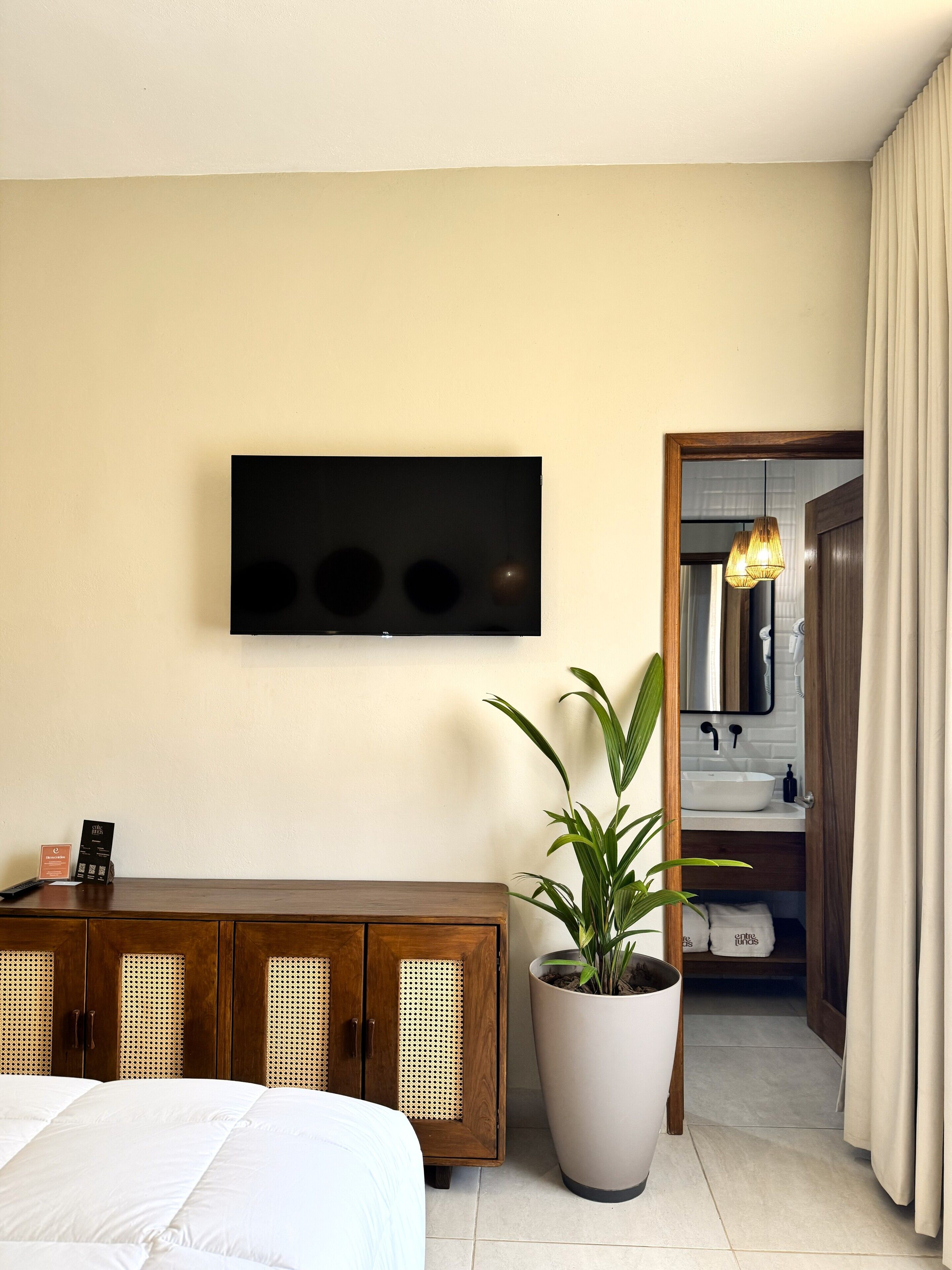Habitación de hotel con una cama, un mueble de madera, una planta en maceta, un televisor de pantalla plana montado en la pared y un baño visible a través de una puerta abierta.
