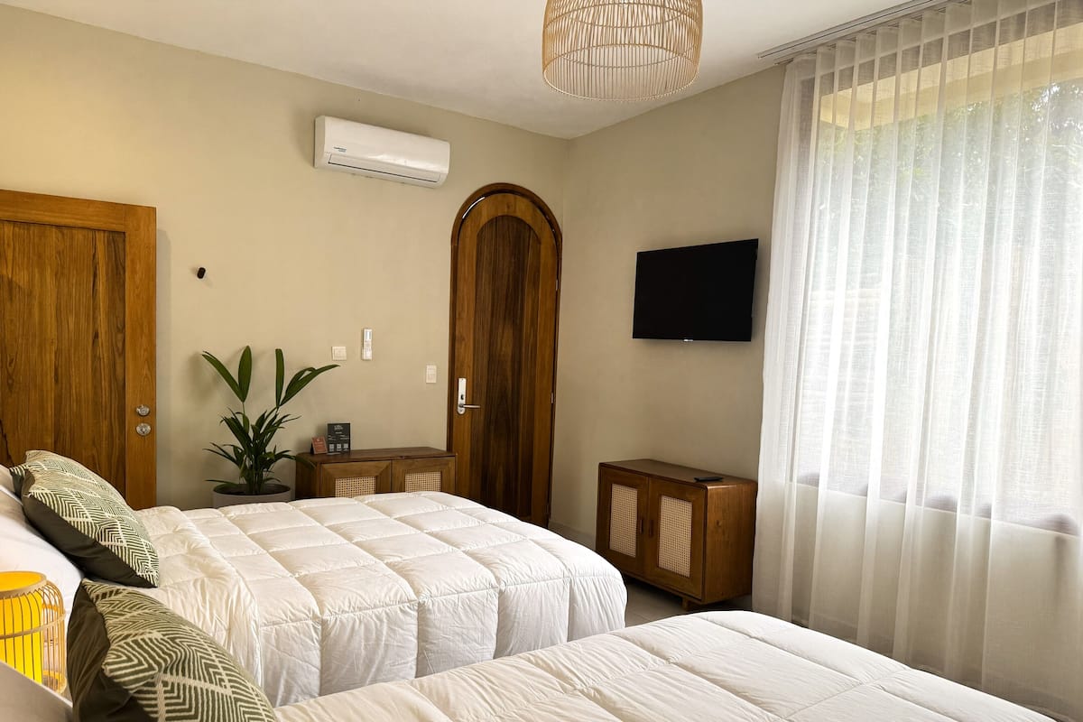 A hotel room with two beds, an air conditioning unit, a television, a wooden wardrobe, a small cabinet, and a potted plant.
