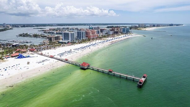 Une vue aérienne d’une ville côtière, avec une longue jetée qui s’avance dans l’eau, une plage parsemée de parasols et, en arrière-plan, des bâtiments et un pont.