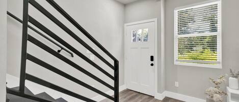 A staircase with a black railing leading to a door, a window with blinds, and a vase with flowers on a pedestal.