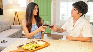 Una pareja sentada en la encimera de la cocina con una tabla de quesos y uvas, sosteniendo copas de vino.