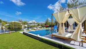 A swimming pool with lounge chairs and a covered seating area, surrounded by well-maintained grass and a body of water.