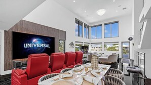 A spacious living room with a large flat-screen TV displaying 'UNIVERSAL', a red sofa, a dining table with a woven placemat, and a view of the outdoors through large windows.
