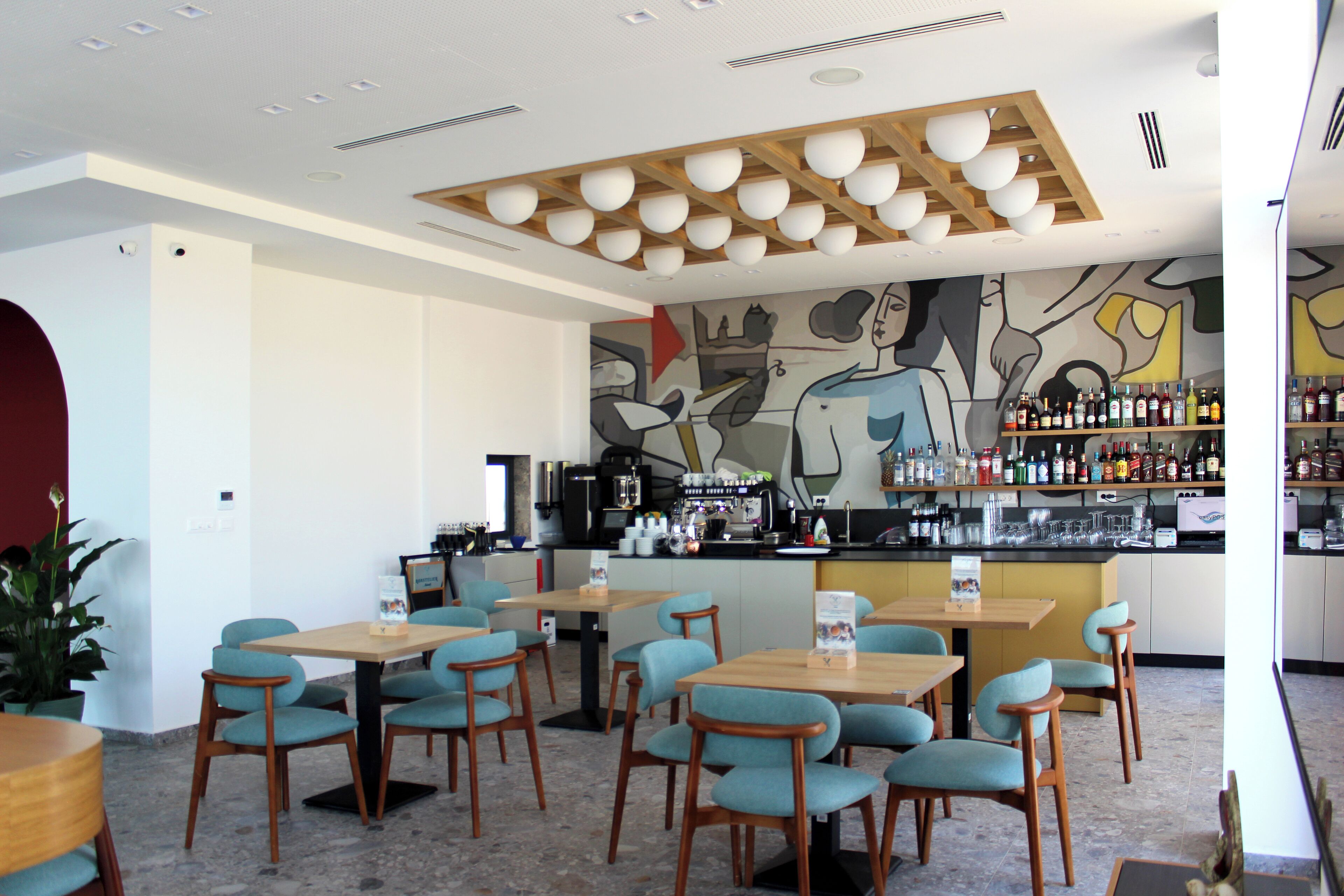 A restaurant with wooden tables and chairs, a bar area with shelves of bottles, and a mural on the wall.