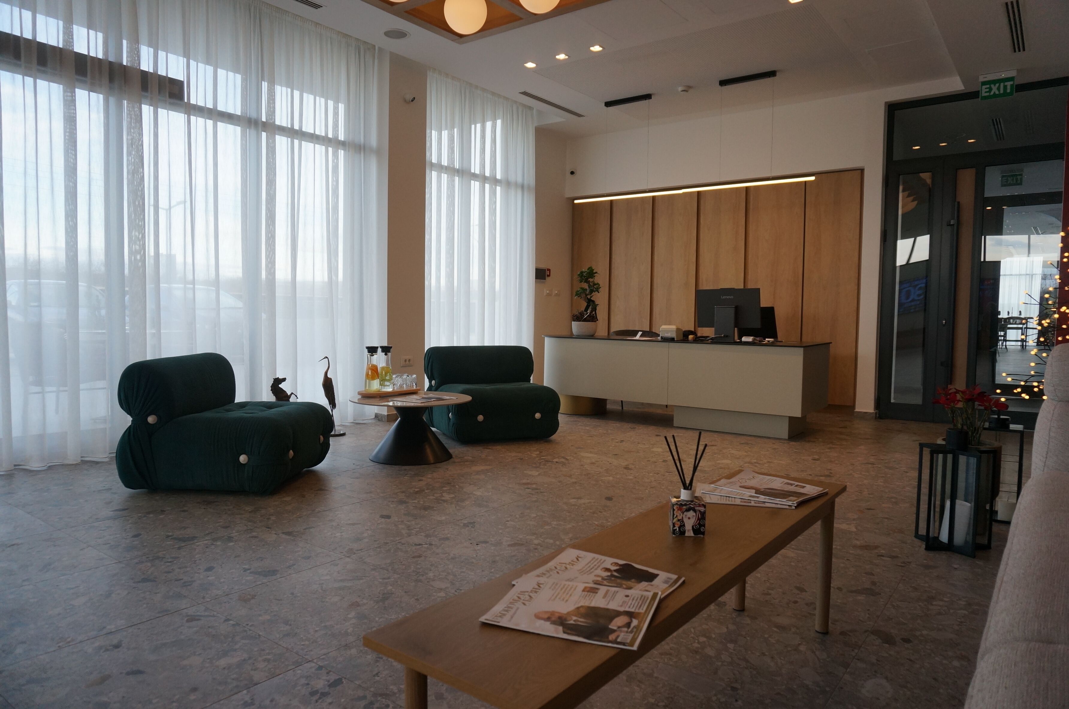 Un moderno vestíbulo de hotel con una recepción, dos sillones verdes, una mesa de centro de madera y una revista sobre un banco de madera.