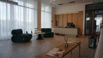 Un moderno vestíbulo de hotel con una recepción, dos sillones verdes, una mesa de centro de madera y una revista sobre un banco de madera.
