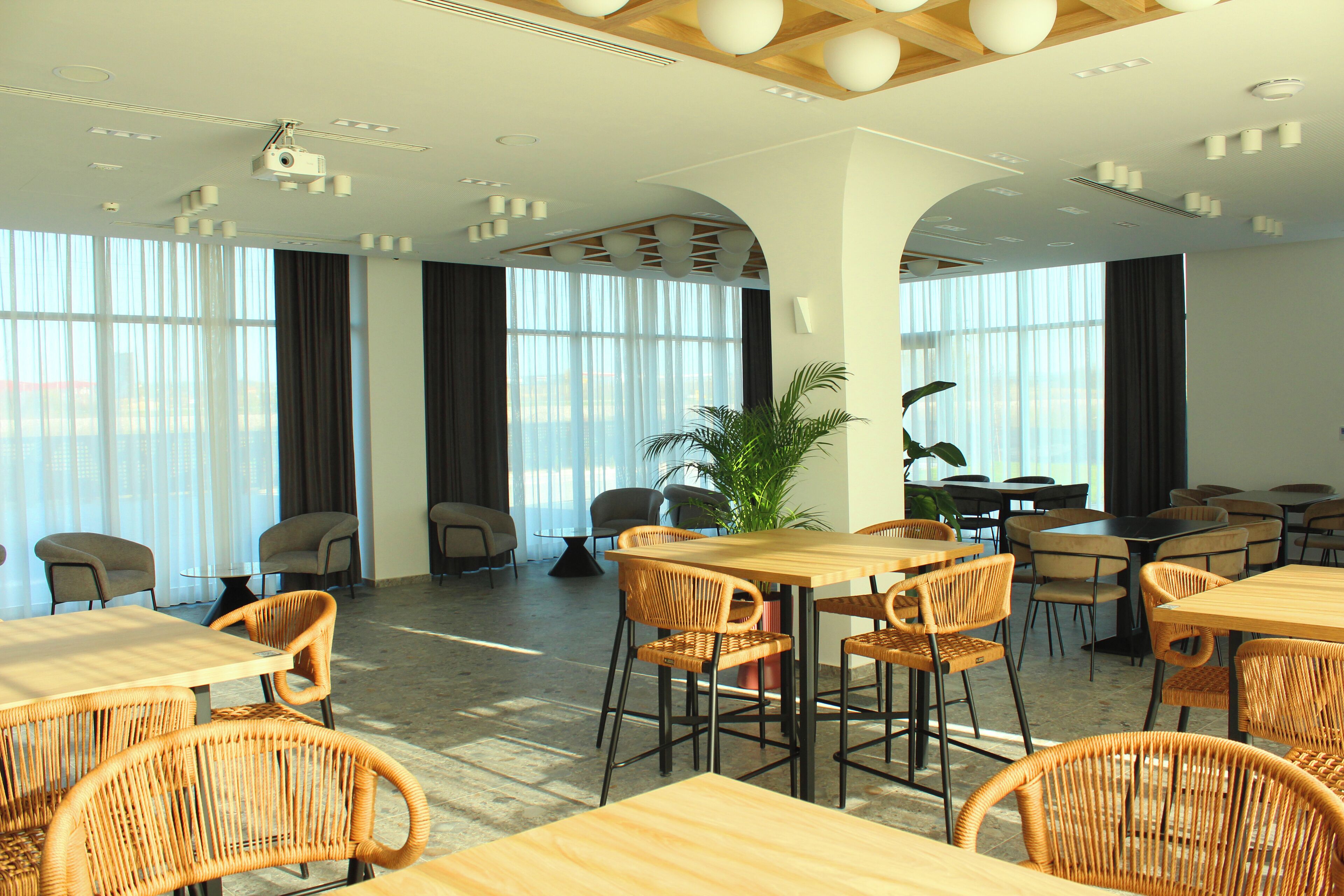 Un comedor moderno con mesas de madera y sillas de mimbre, ventanas amplias con cortinas e iluminación empotrada en el techo.