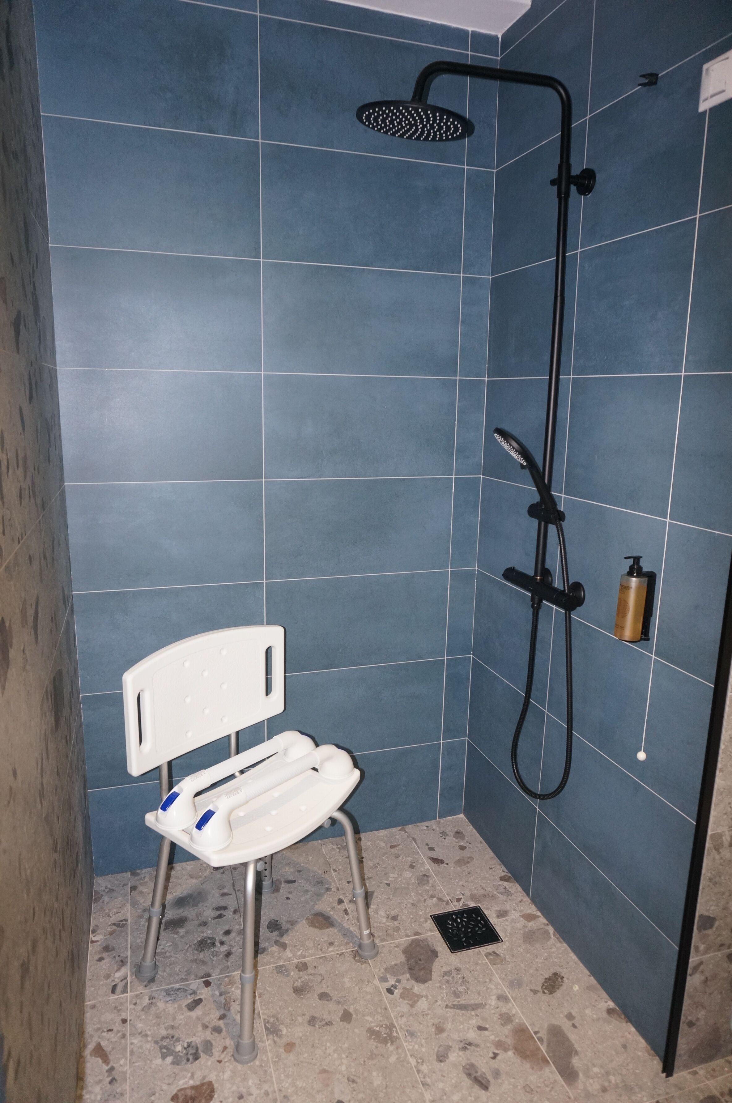 Douchegedeelte met blauwe tegels, een witte douchestoel en een handdouche.