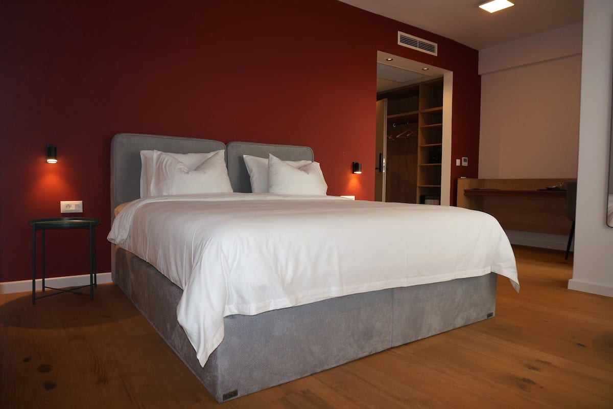Un lit bien fait, avec une tête de lit grise et du linge de lit blanc, dans une pièce au plancher en bois et aux murs rouges.