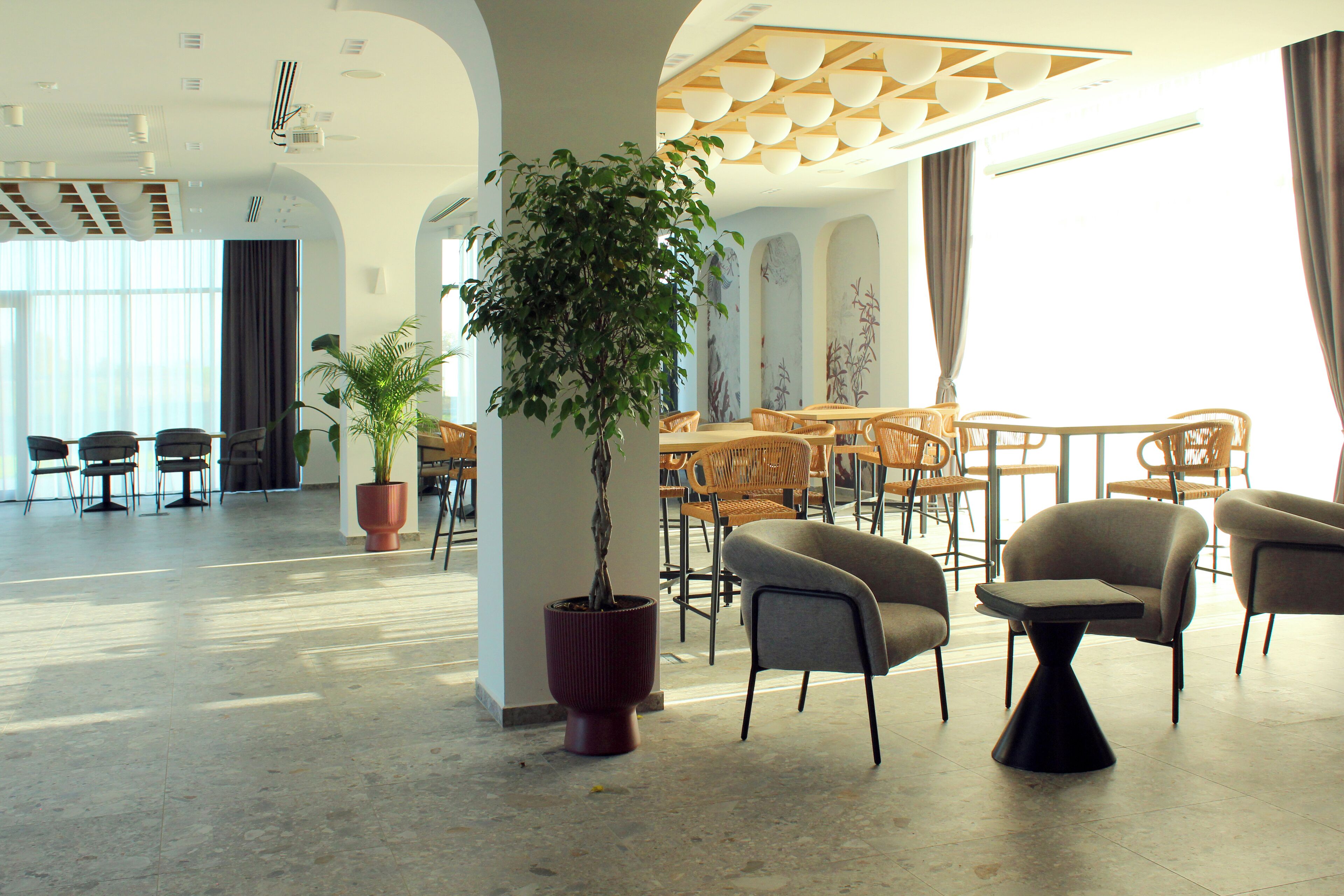 A spacious lobby with a modern design, featuring a large potted plant, comfortable seating areas, and a high ceiling with unique lighting fixtures.